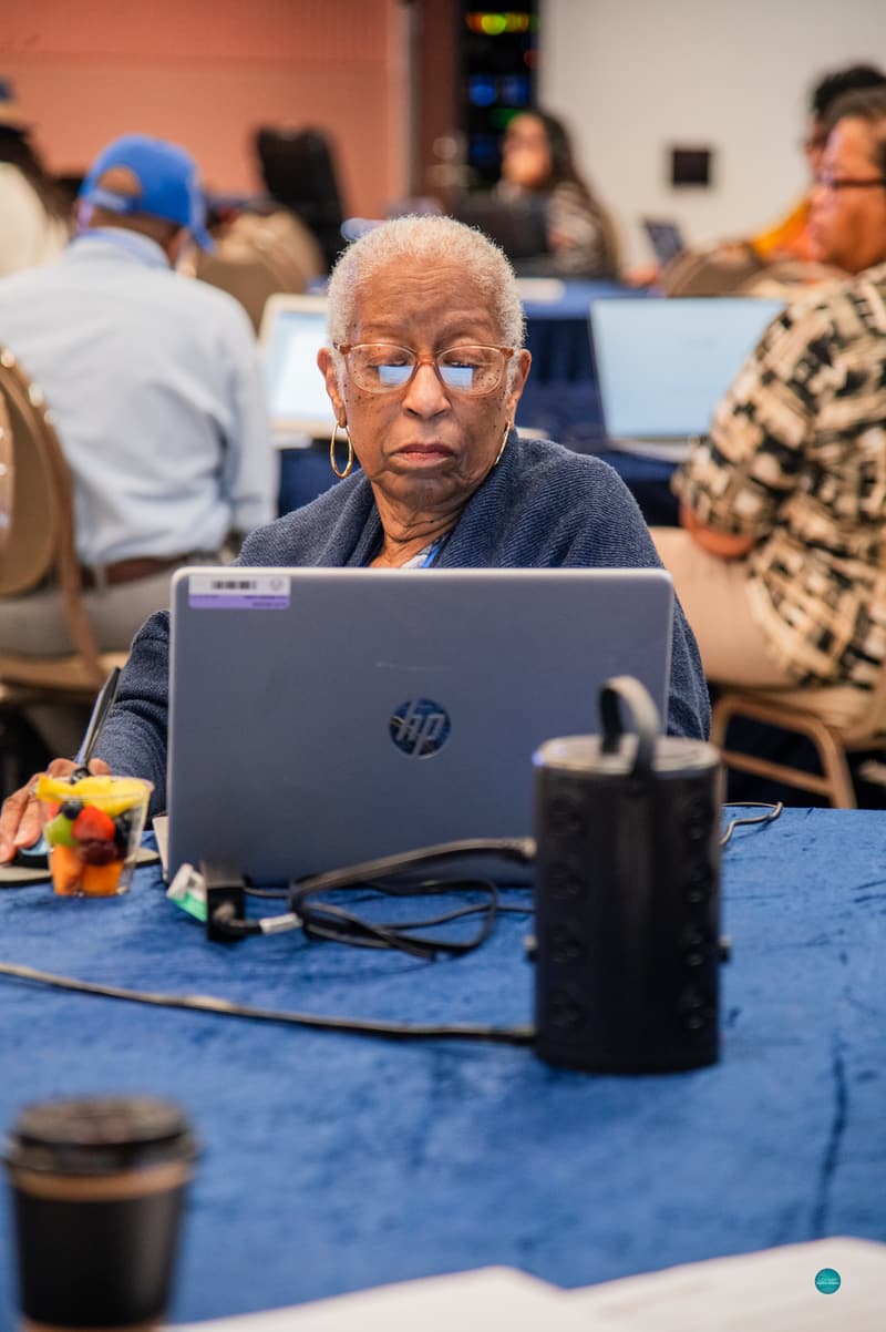 An older attendee with short gray hair, glasses, and gold hoop earrings works on a laptop at a blue-covered table, with a cup of fruit beside her and other attendees on laptops in the background.