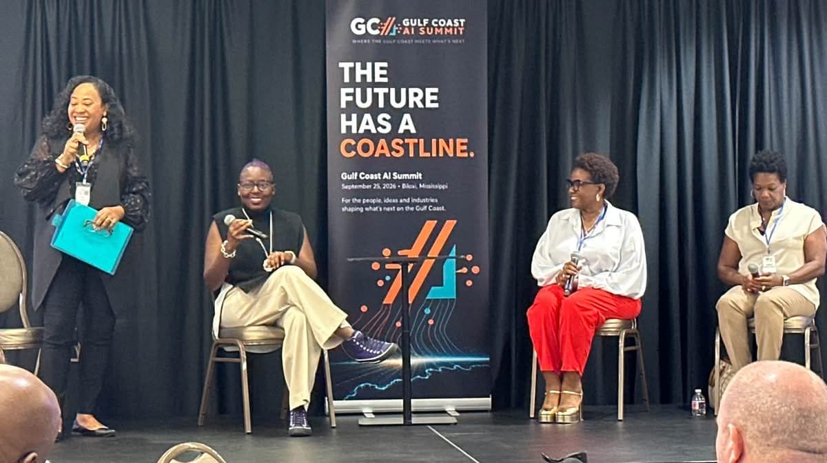 An onstage conversation at the Summit: a moderator standing with a microphone at left and three seated panelists holding microphones, beside a banner reading &ldquo;The Future Has a Coastline. Gulf Coast AI Summit, September 25, 2026, Biloxi, Mississippi.&rdquo;
