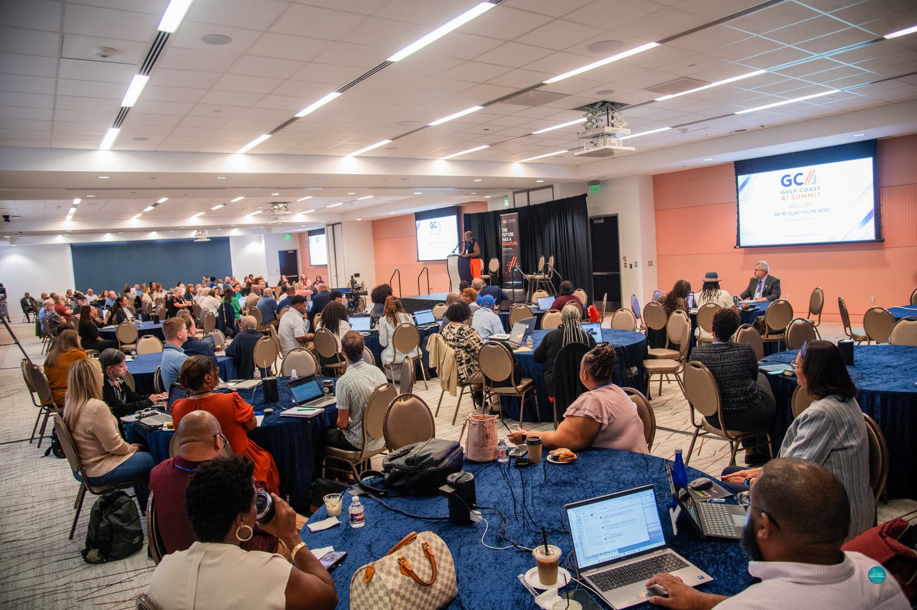 The Summit room during the program: attendees with laptops and coffee at round tables with blue tablecloths face a speaker at a podium on stage, with a large screen reading &ldquo;Gulf Coast AI Summit &mdash; Welcome, we&rsquo;re glad you&rsquo;re here.&rdquo;