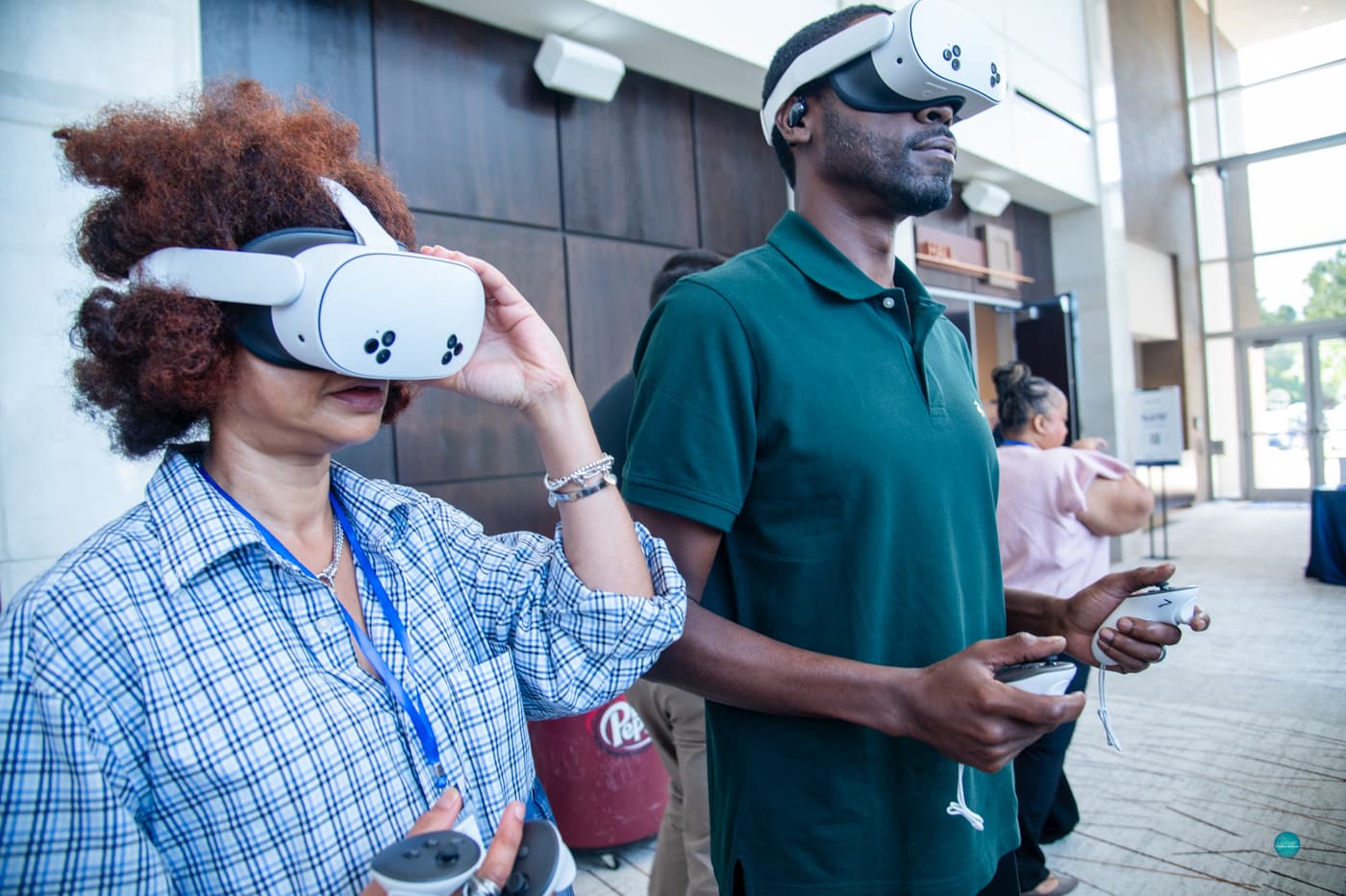 Two attendees try the AR/VR demonstration in the venue lobby: a woman in a plaid shirt and Summit lanyard adjusts a white VR headset while a man in a green polo stands beside her wearing a headset and holding both controllers.